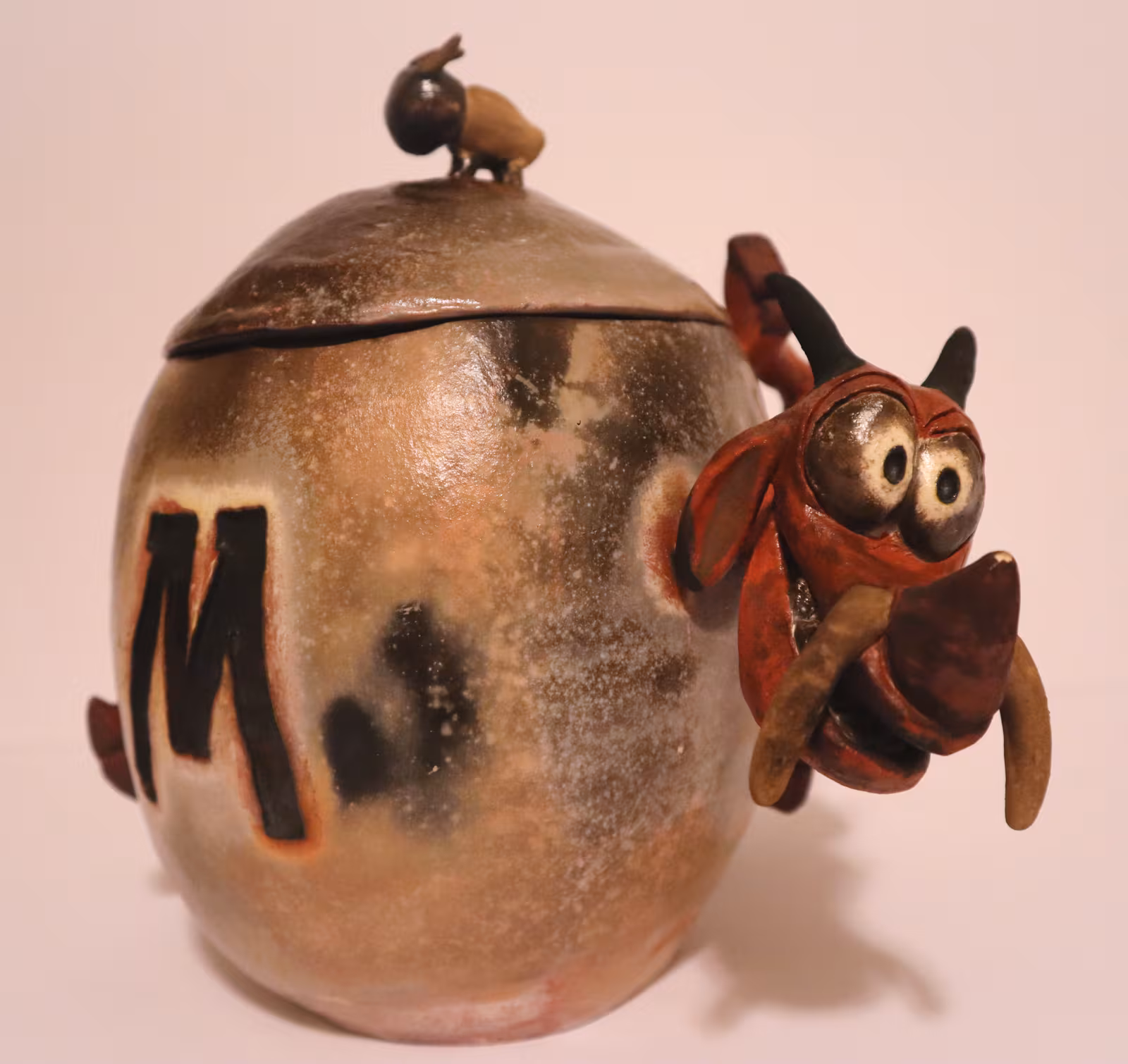 A wood-fired lidded ceramic jar marked with an M, a small bird on the lid and a red clay creature with big eyes clinging to the side.