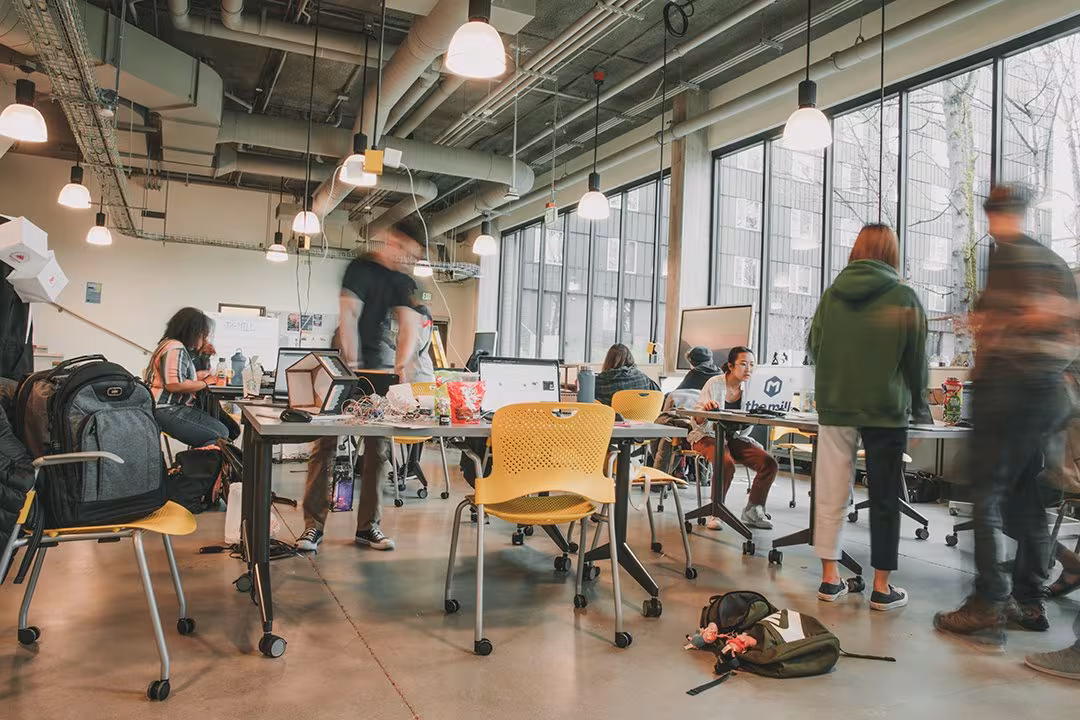 An open prototyping studio with students at long tables, laptops, electronics and half-built models, and tall windows along one wall